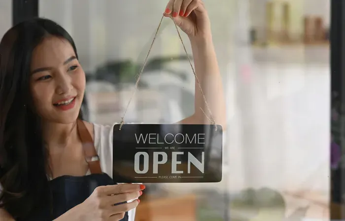 Smiling shop owner hanging a welcome open sign on the front door, inviting customers to visit and discover local services